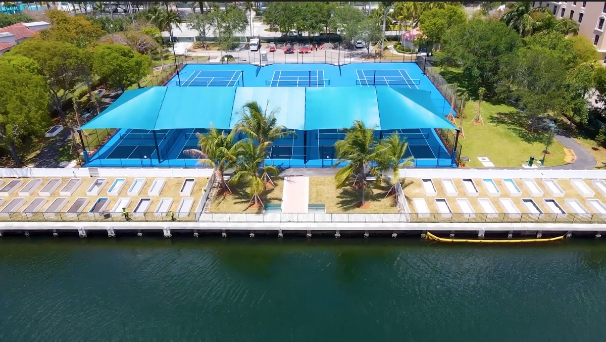 Aerial view of an outdoor swimming pool complex with a blue canopy cover next to a waterfront dock or marina. White lounge chairs line the pool deck and palm trees provide landscaping.