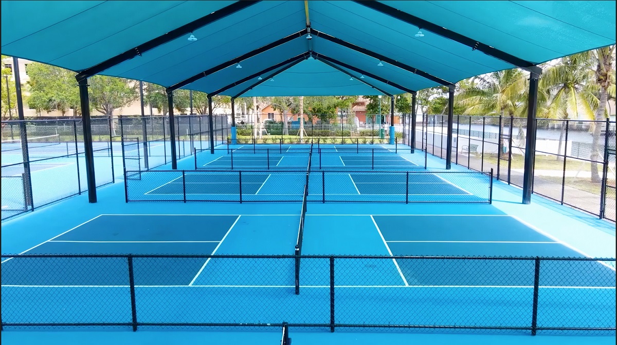 A covered outdoor swimming pool with multiple lanes divided by lane lines, featuring a blue canopy roof and surrounded by a fence. The pool appears empty and well-maintained.