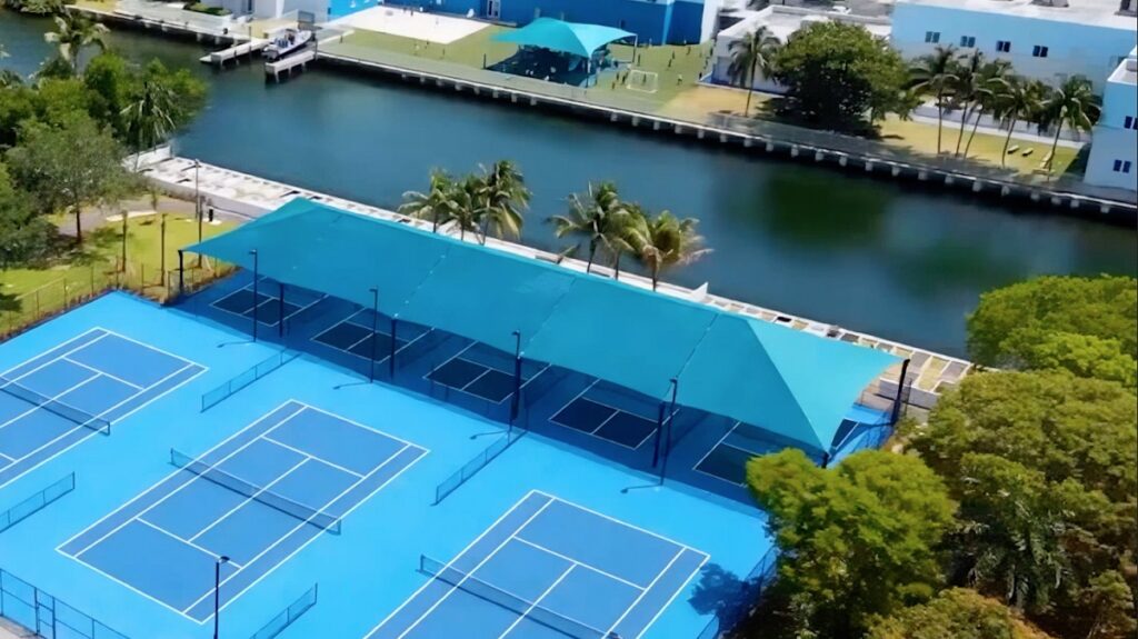 Aerial view of bright blue tennis courts next to a waterway, with palm trees and boats visible along the water's edge.